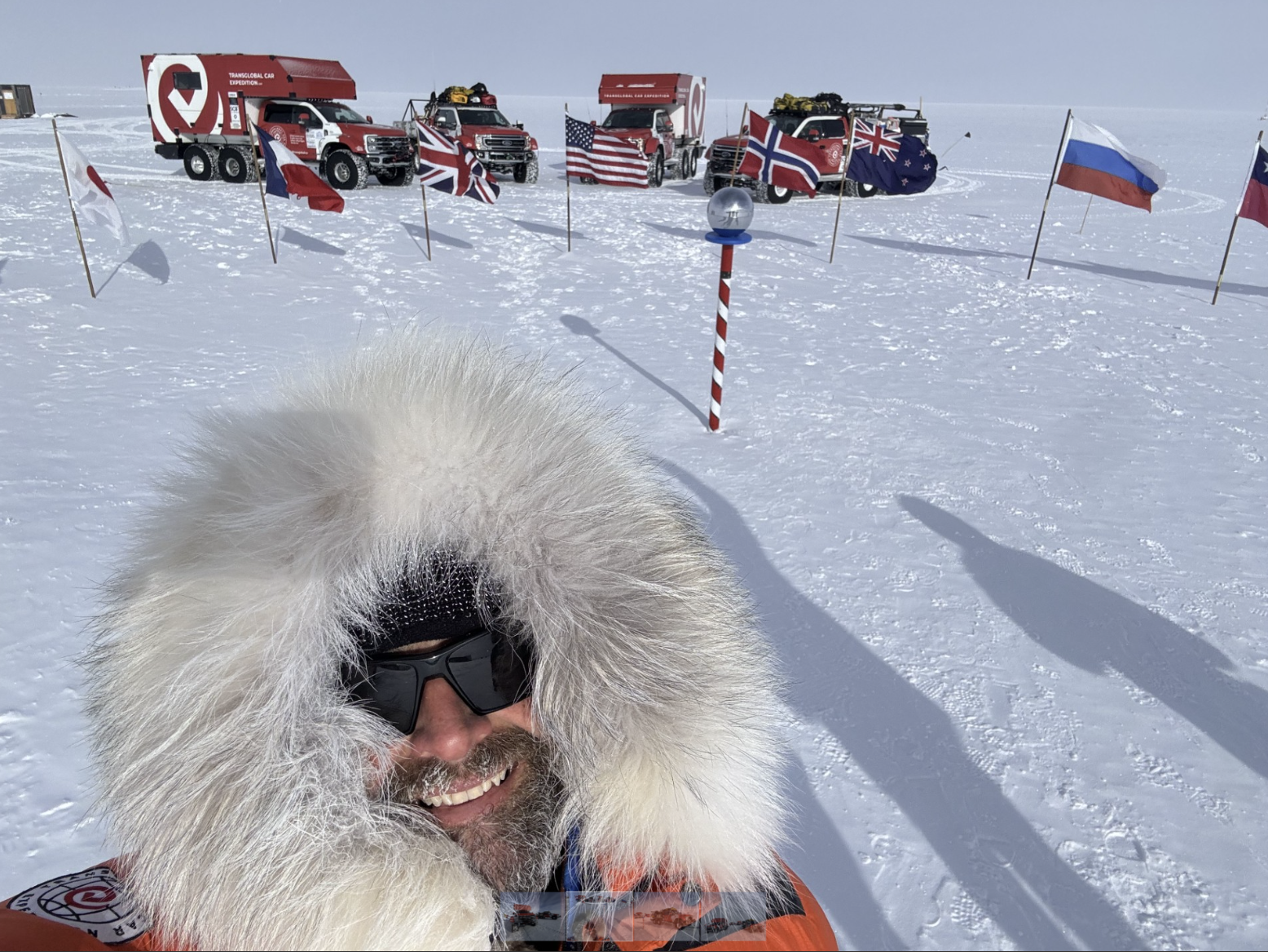 A man wearing sunglasses and a coat with a fur-covered hood stands in front of red Ford vehicles at the South Pole. 